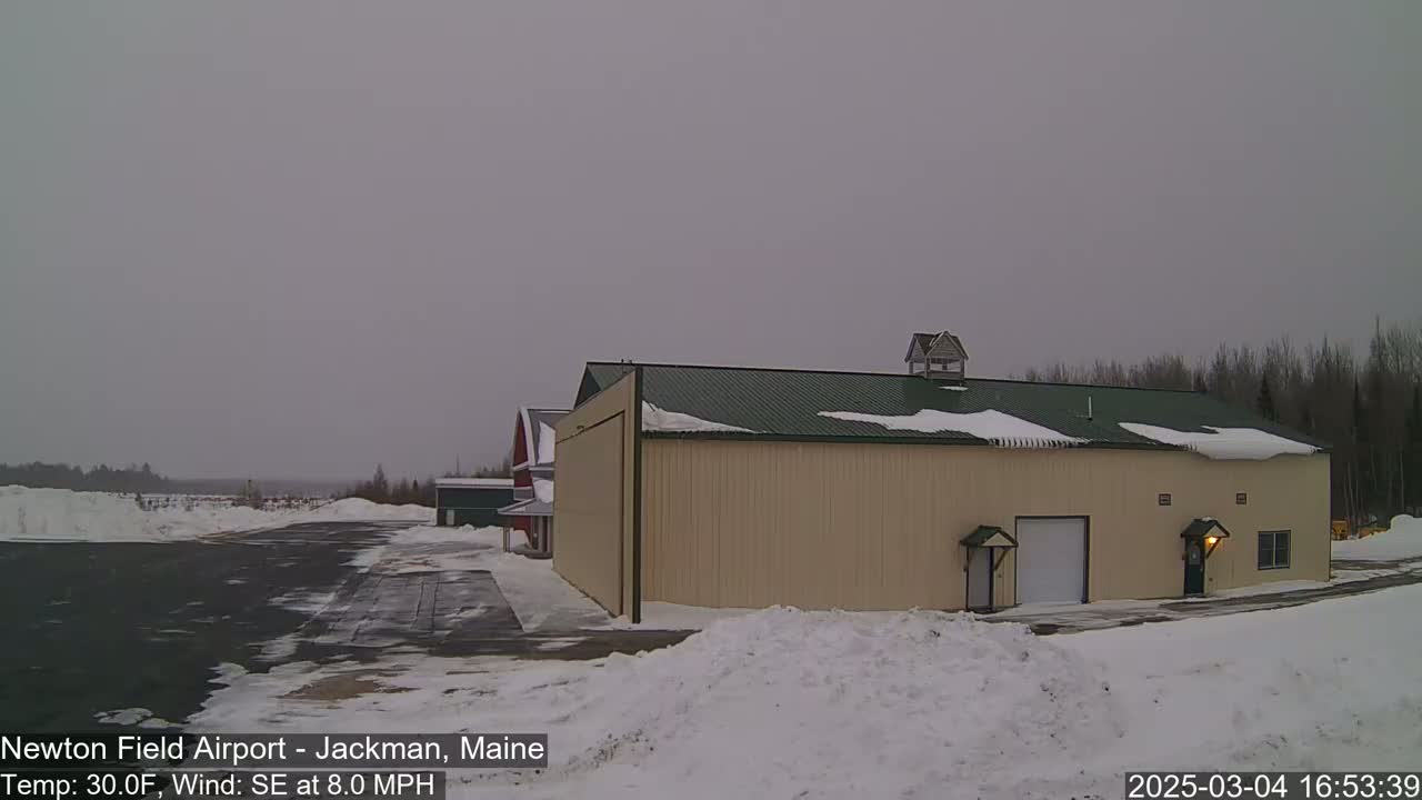A large tan hangar with a green roof sits on a snowy airfield under an overcast sky.