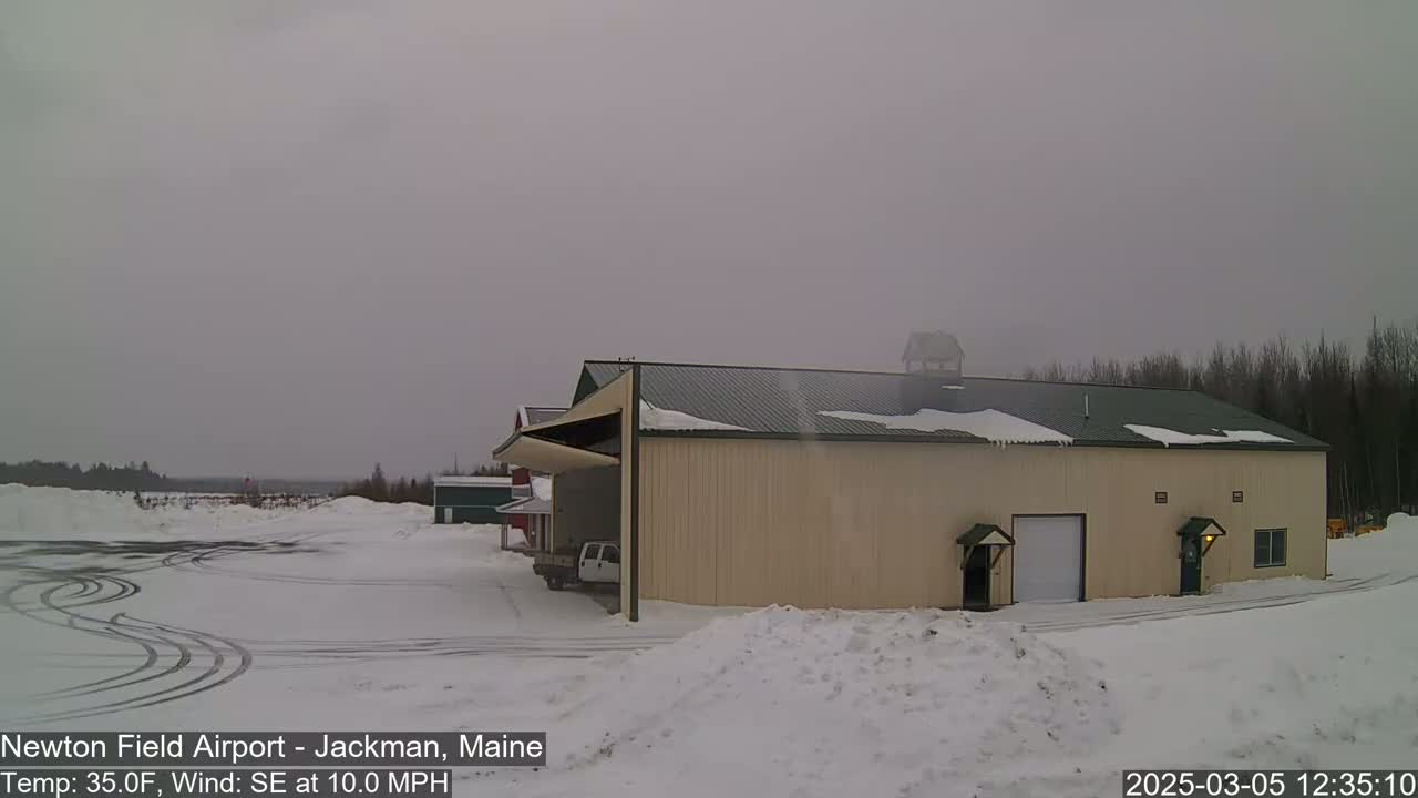 A large tan building with a green roof sits in a snowy landscape under an overcast sky, with a pickup truck parked nearby.