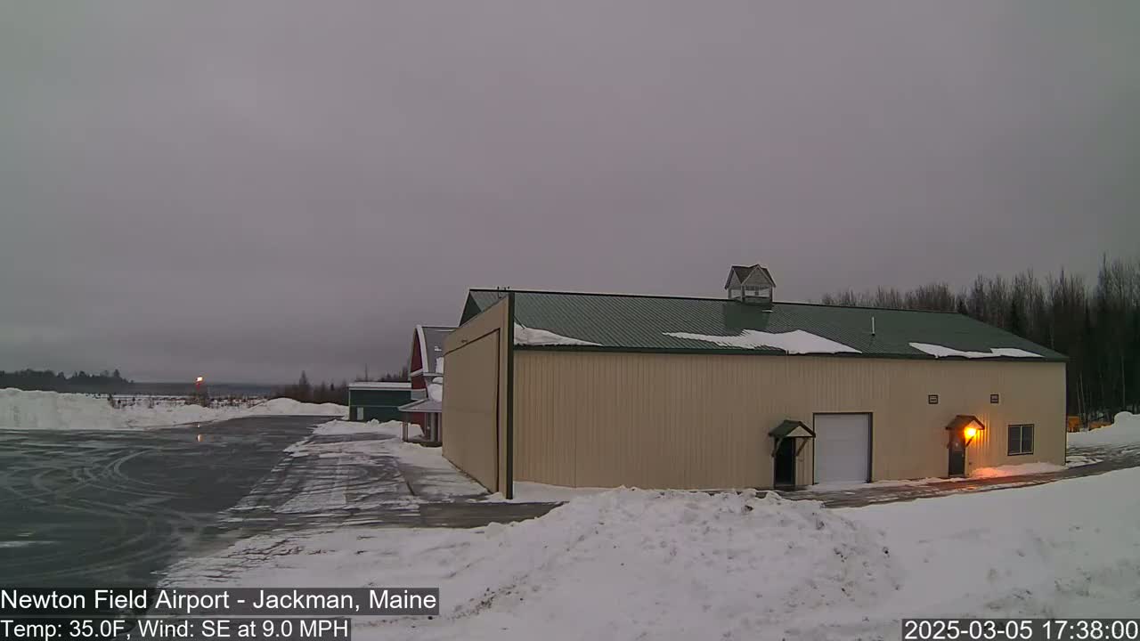 A tan hangar with a green roof sits on a snow-covered airfield under an overcast sky.