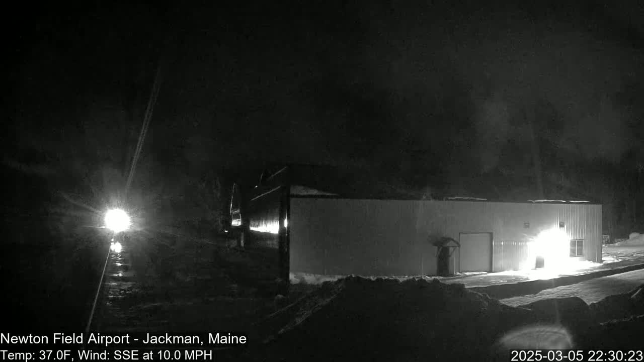 A long, single-story building is illuminated at night in snowy conditions, with headlights visible in the distance.