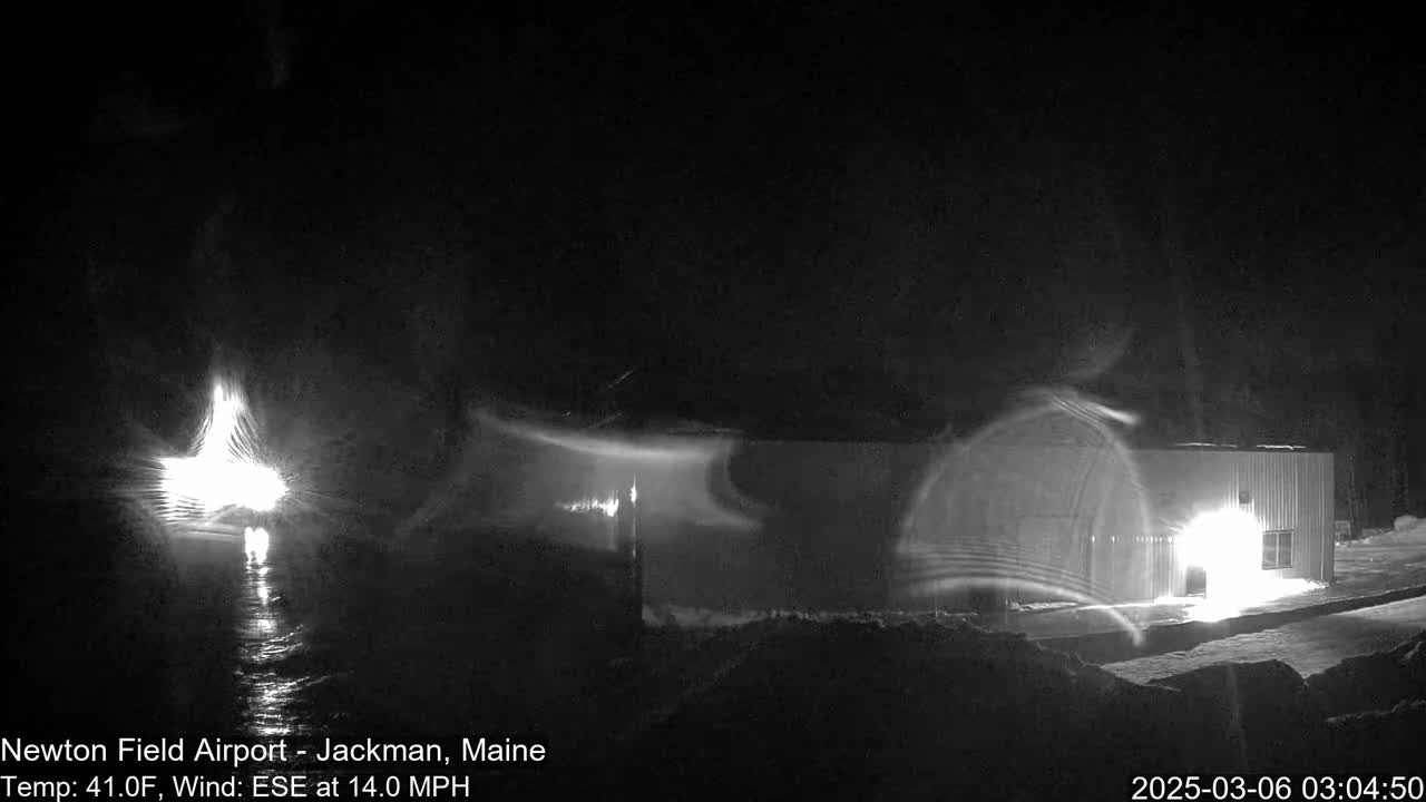 A nighttime view of a building illuminated by lights, with strong winds causing snow or blowing debris to swirl around it.