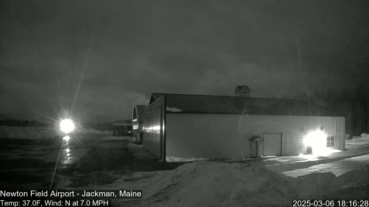 A large hangar-like building is illuminated at night in snowy conditions under a dark, cloudy sky.