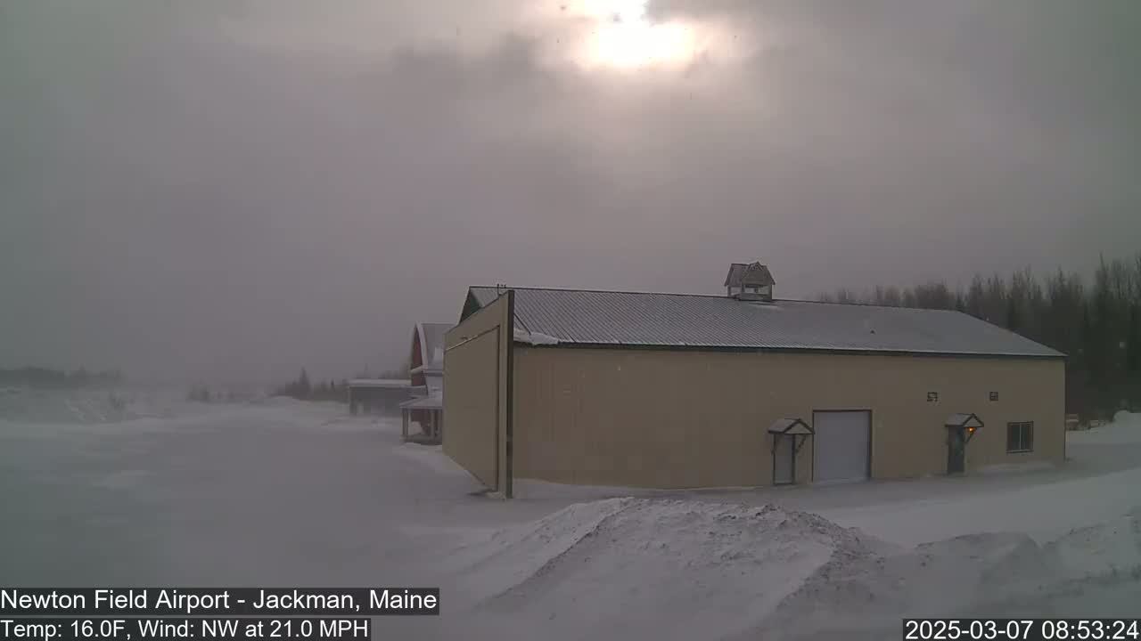 A tan hangar sits in a snowy landscape under an overcast sky, with a faint sun visible and a strong northwest wind.