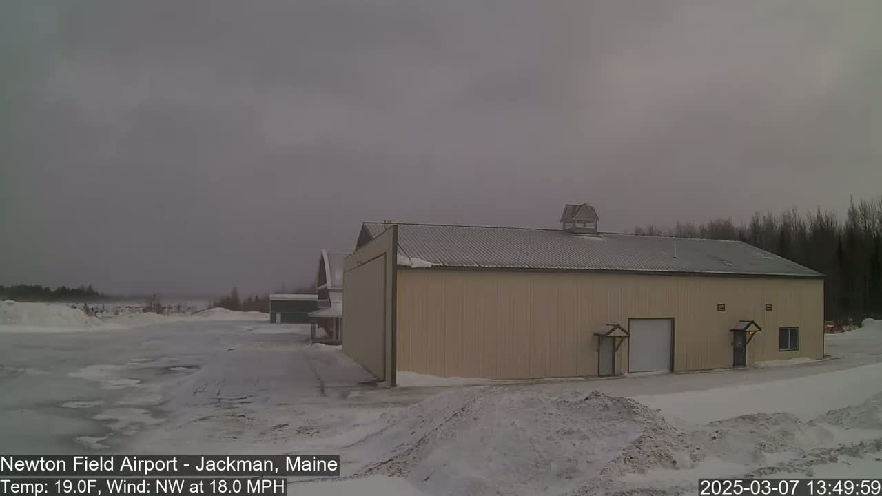 A large tan hangar sits in a snow-covered airport under a gray, overcast sky with a northwest wind blowing at 18 mph.