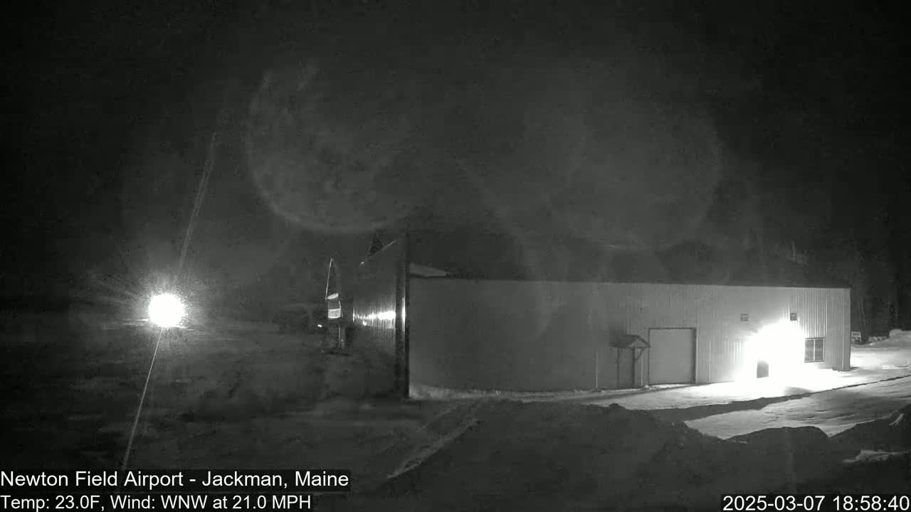 A long, low building is illuminated at night in snowy conditions with a strong wind.