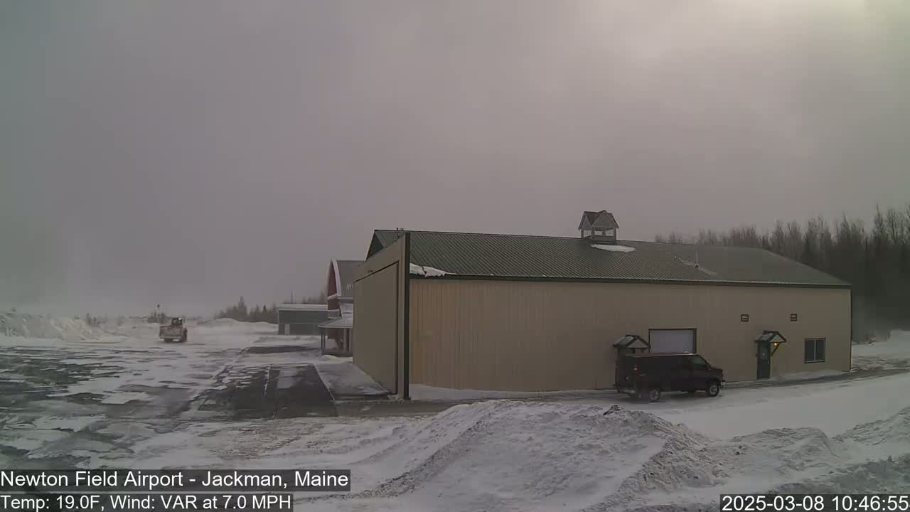 A large tan hangar sits in a snowy airfield with a dark-colored van parked in front of it under an overcast sky.