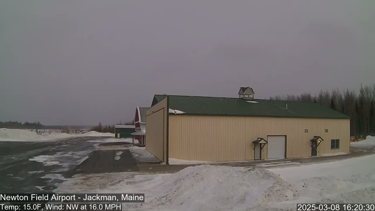 A large tan hangar with a green roof sits on a snowy airfield under an overcast sky.