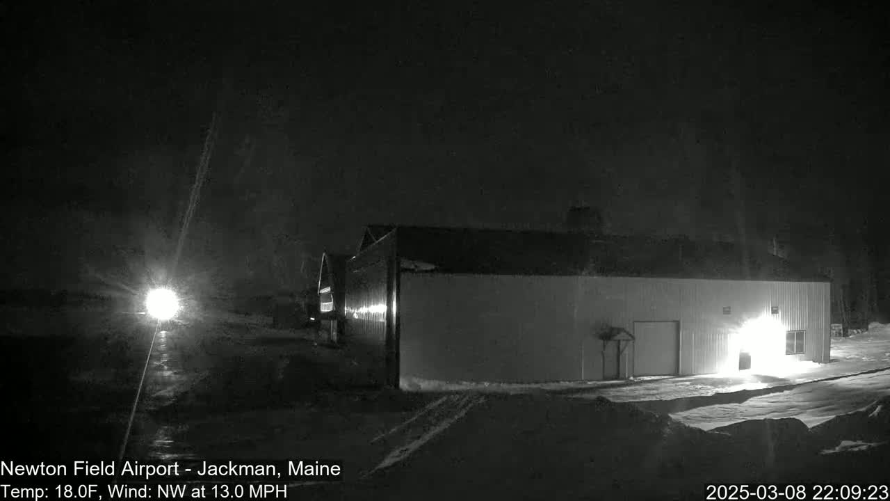 A large, light-colored building is illuminated at night in snowy conditions with a northwest wind blowing at 13 mph and a temperature of 18°F.