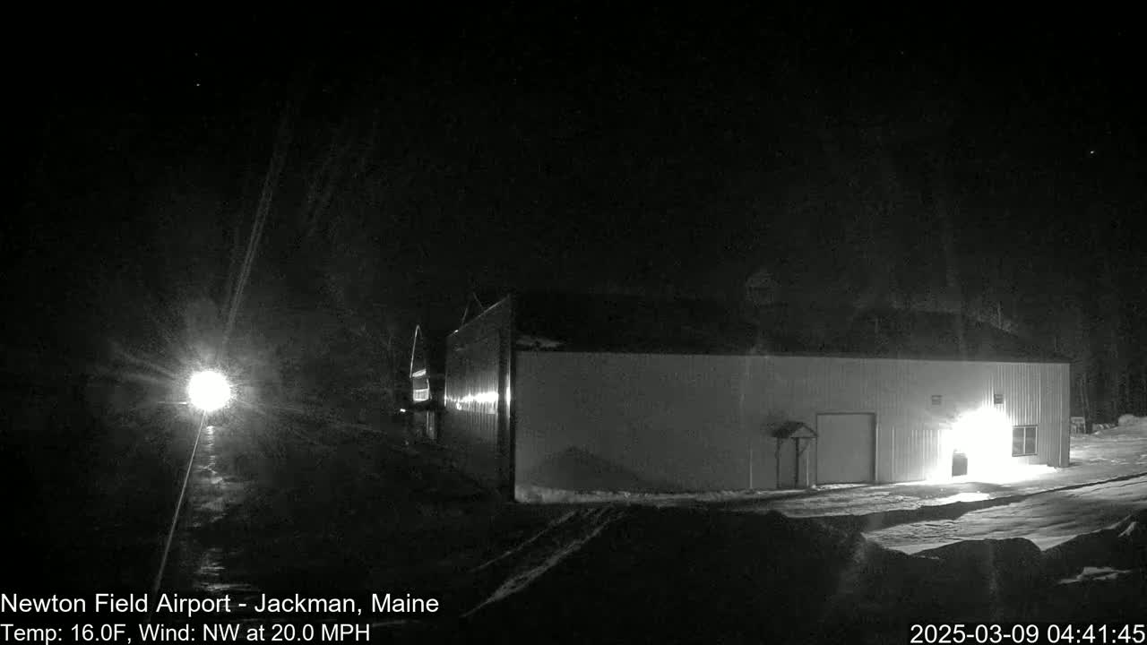 A large, light-colored building is illuminated at night in snowy conditions with a strong northwest wind.