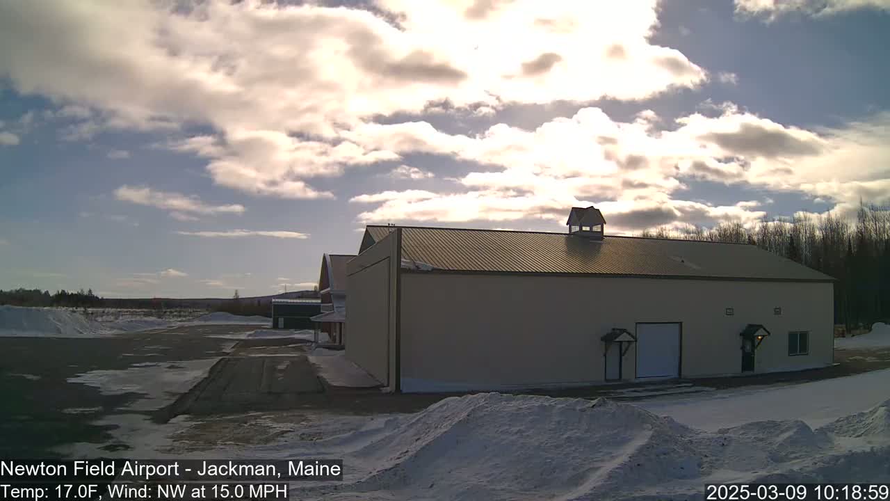 A large tan building with a metal roof sits in a snowy airfield under a partly cloudy sky.