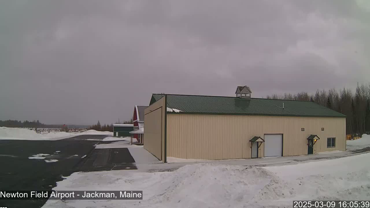 A large tan hangar with a green roof stands in a snowy airfield under an overcast sky.