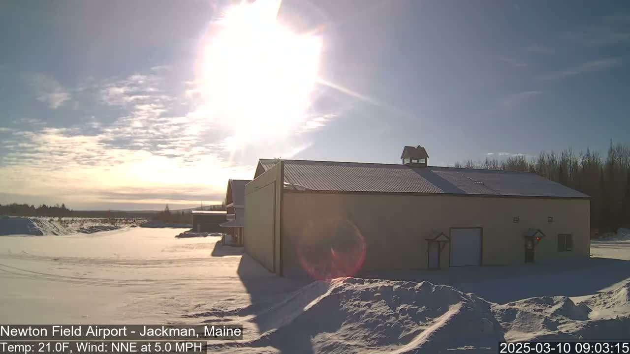 A tan building with a snow-covered roof stands in a snowy landscape under a bright, sunny sky with scattered clouds.