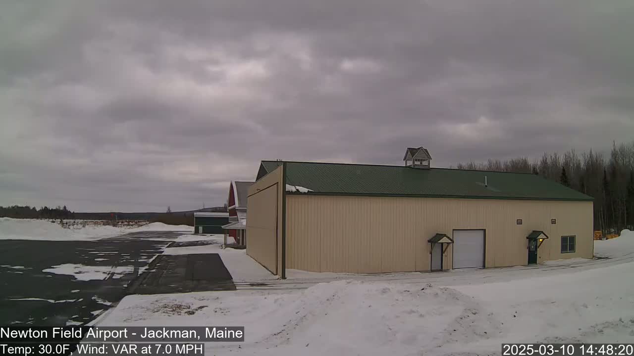 A large tan hangar with a green roof sits on a snowy airfield under an overcast sky.
