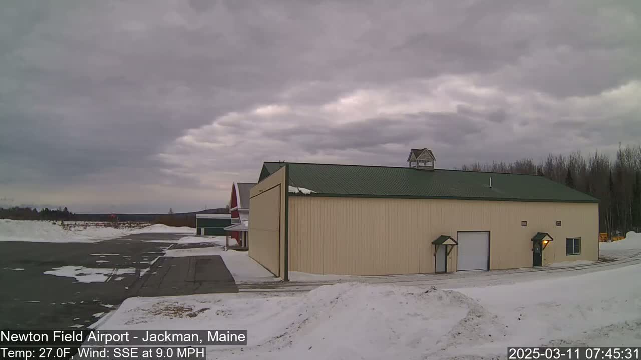 A large tan hangar with a green roof sits on a snowy airfield under an overcast sky.