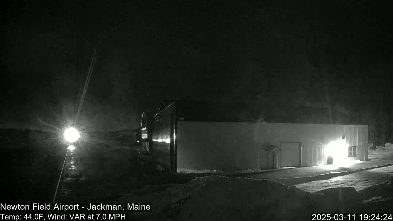 A long, low building is illuminated at night in snowy conditions, with a bright light source visible in the distance.