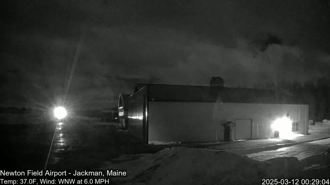 A large, dark building is illuminated at night in snowy conditions with a bright light source visible in the distance.