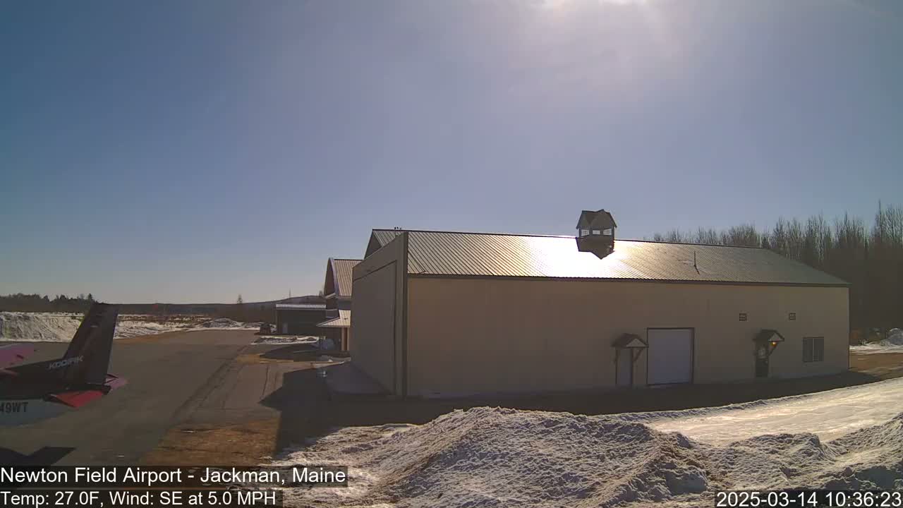 A small airplane is parked on an airfield next to a tan building on a sunny day with snow on the ground.