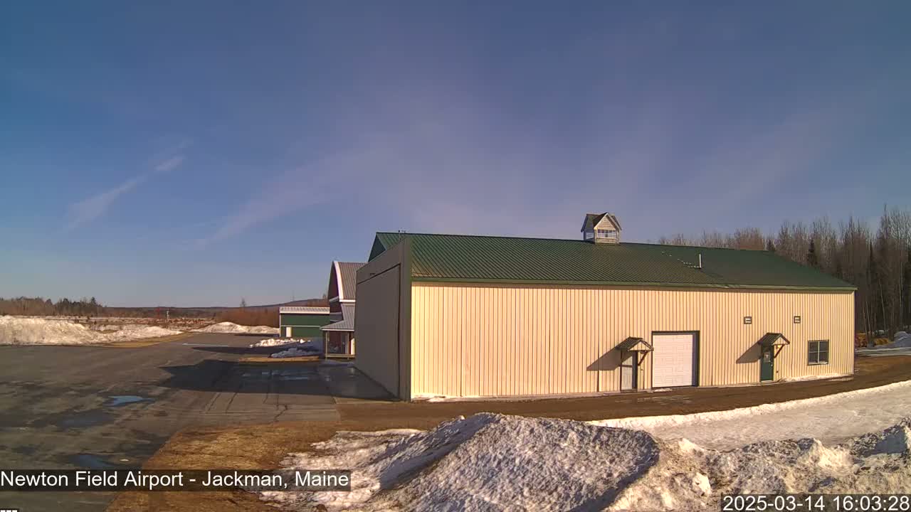 A large tan hangar with a green roof sits on an airfield on a sunny day with patches of snow on the ground.