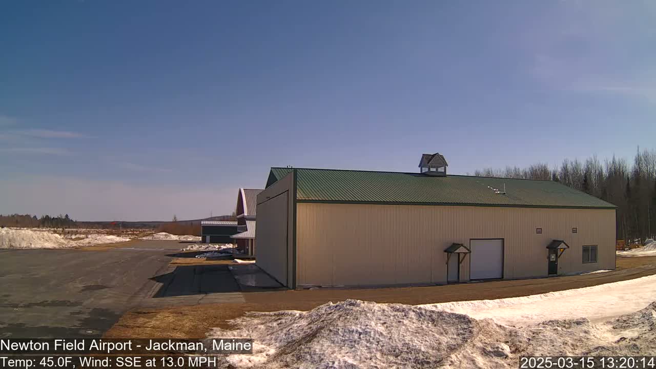 A large tan building with a green roof sits on a mostly cleared airfield with patches of snow under a clear blue sky.