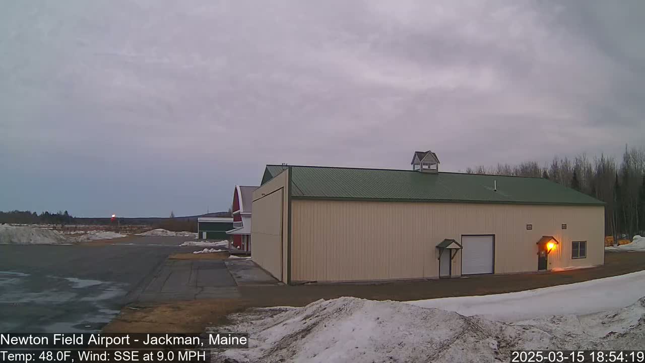 A large tan hangar with a green roof stands on an airfield under an overcast sky, with patches of snow on the ground.