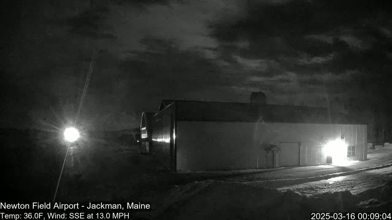 A large building is illuminated at night under a cloudy sky, with snow on the ground and a light source visible in the distance.