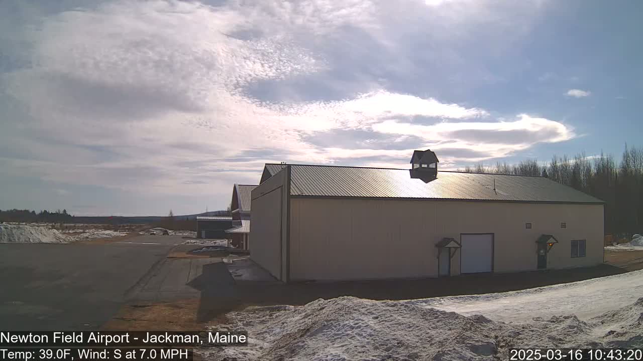 A large tan building with a green roof sits on a mostly clear day with a light south wind, next to an airport runway with patches of snow on the ground.