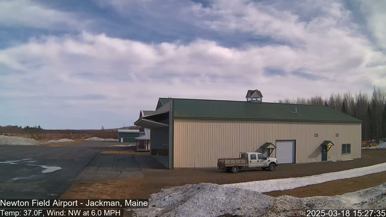 A large beige hangar with a green roof and a pickup truck parked in front sits on an airfield under a partly cloudy sky.