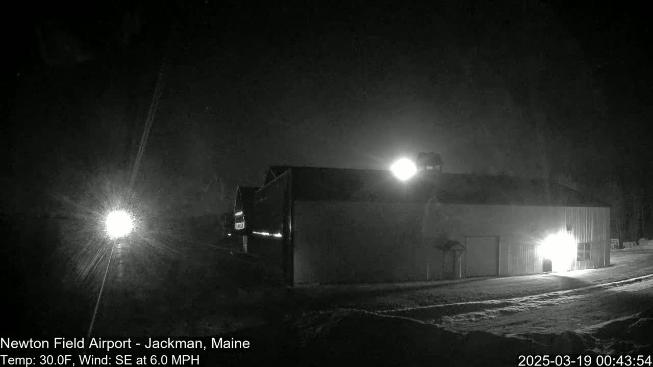 A large, light-colored building is illuminated at night in snowy conditions, with a bright light source visible in the distance.
