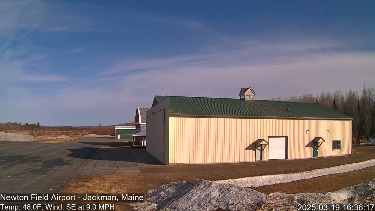 A large tan hangar with a green roof sits on an airfield under a clear blue sky with a few wispy clouds.