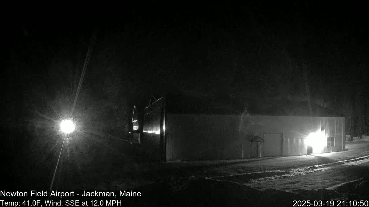 A long, low building is illuminated at night in snowy conditions with a bright light source in the distance.