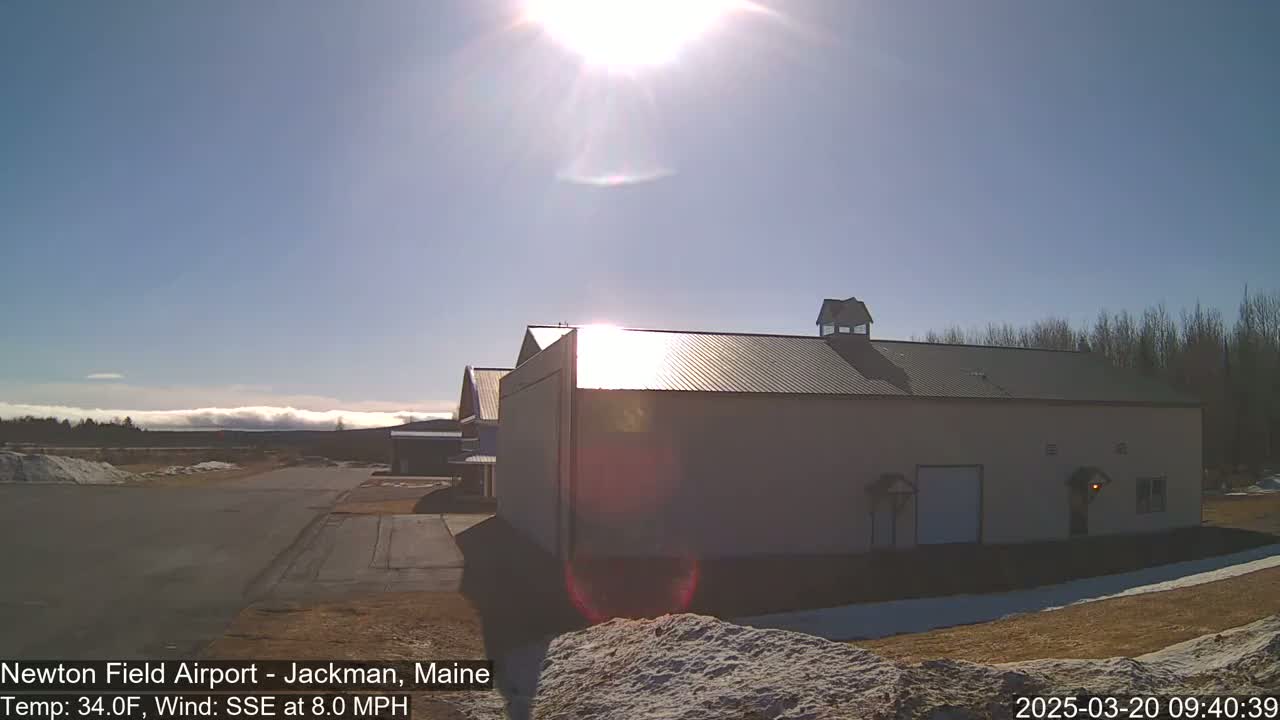 A large tan building with a metal roof sits on a mostly clear, sunny day with a light wind, in front of a distant line of low clouds and bare trees.