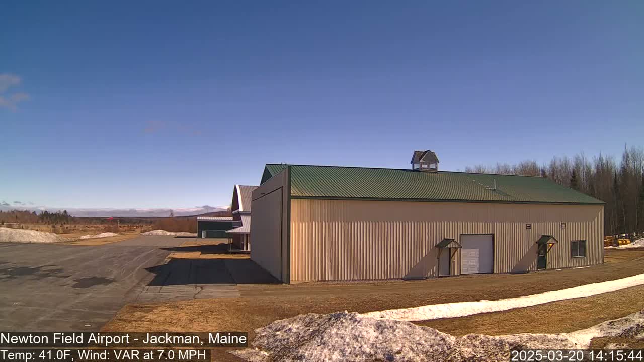 A large tan hangar with a green roof sits on an airfield under a clear blue sky, with patches of snow on the ground.