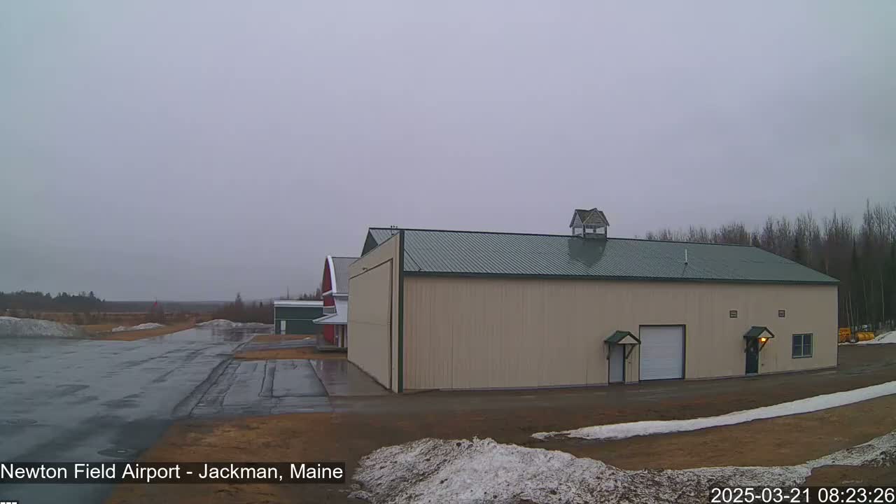A large tan hangar with a green roof sits next to a smaller red barn on a wet, overcast day at an airfield with patches of snow on the ground.