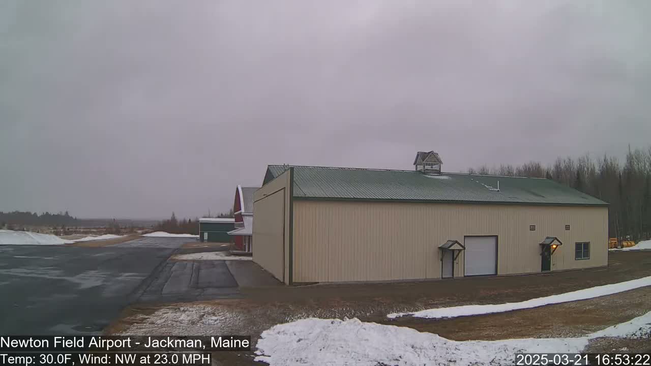 A large tan hangar with a green roof sits near a red barn and a snow-covered airfield under an overcast sky, with a northwest wind at 23 mph and a temperature of 30 degrees Fahrenheit.