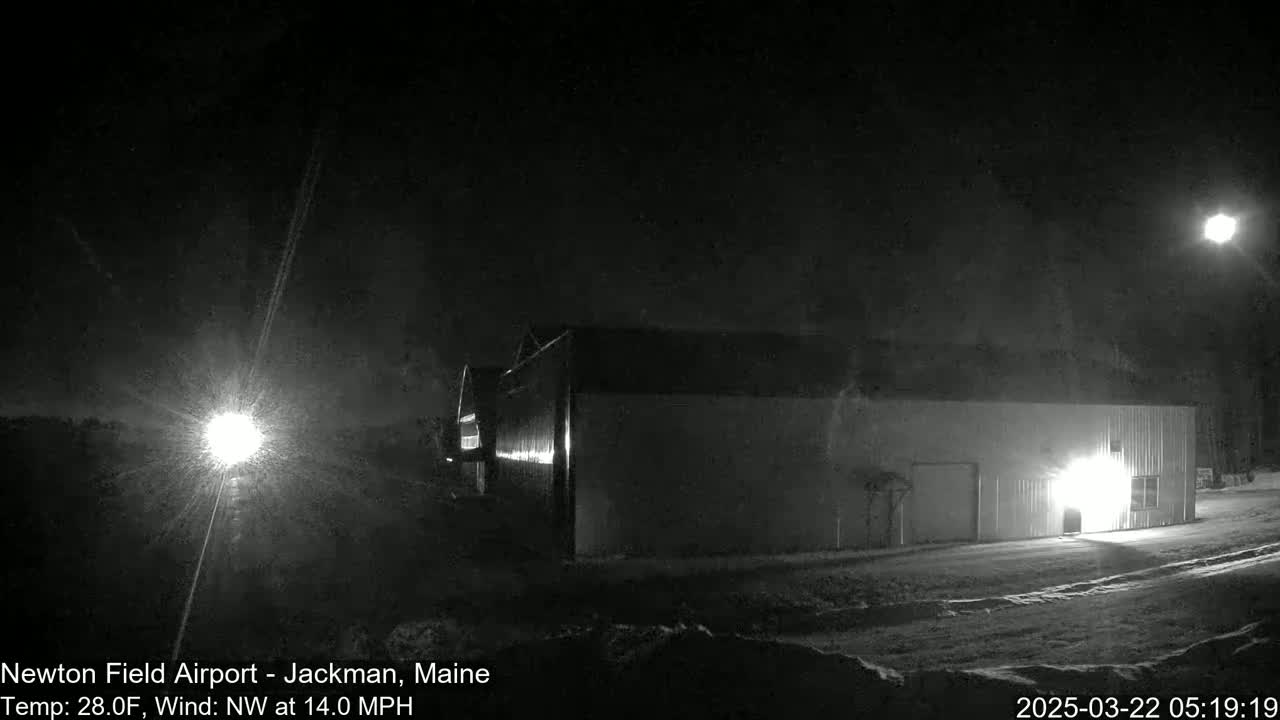 A long, dark building is illuminated by lights at night in snowy conditions with a bright light source in the distance and a Northwest wind of 14 mph.