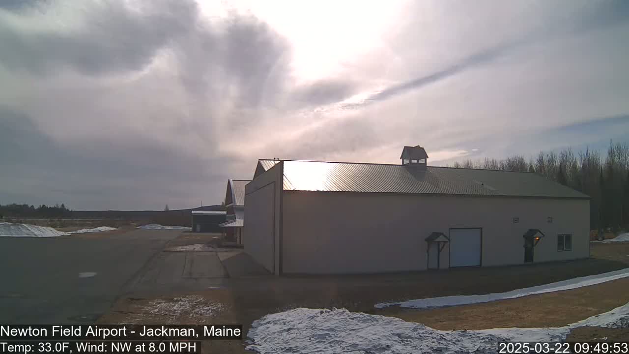 A large, light-colored building with a metal roof sits on an airfield under a partly cloudy sky, with some snow on the ground.