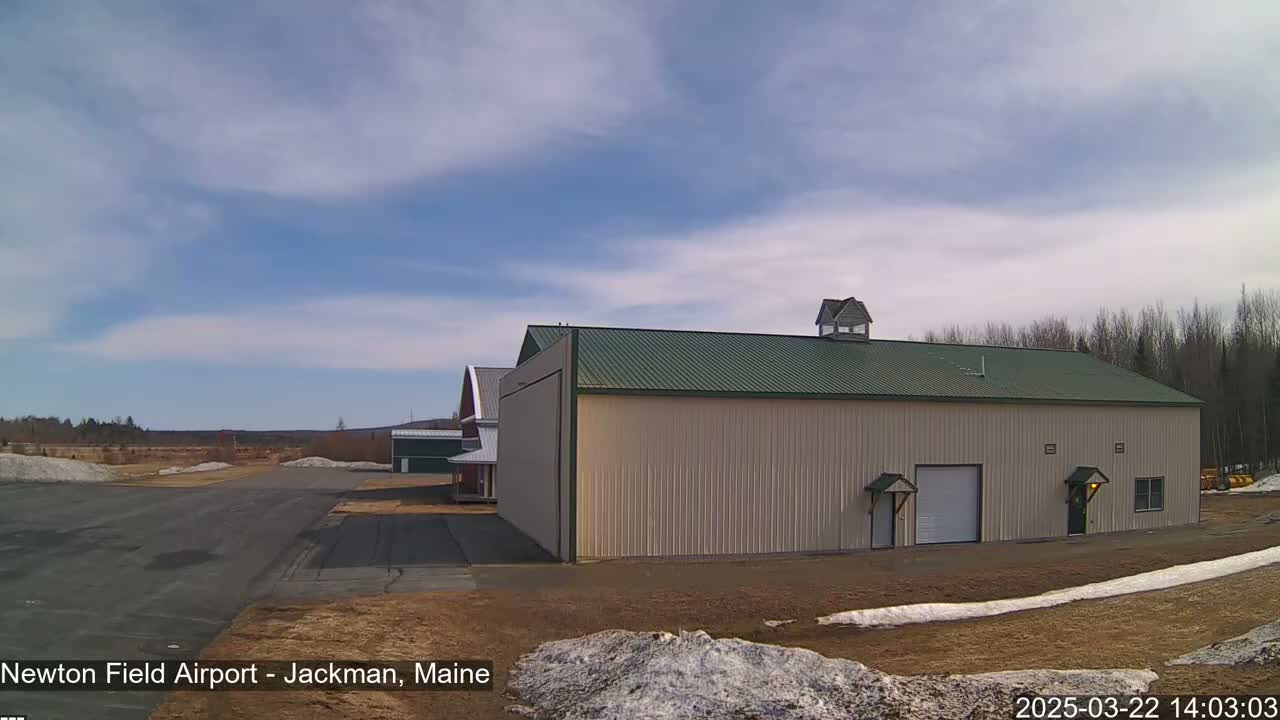 A large tan hangar with a green roof sits on an airport tarmac under a mostly clear, bright sky with a few scattered clouds, and patches of snow remain on the ground.