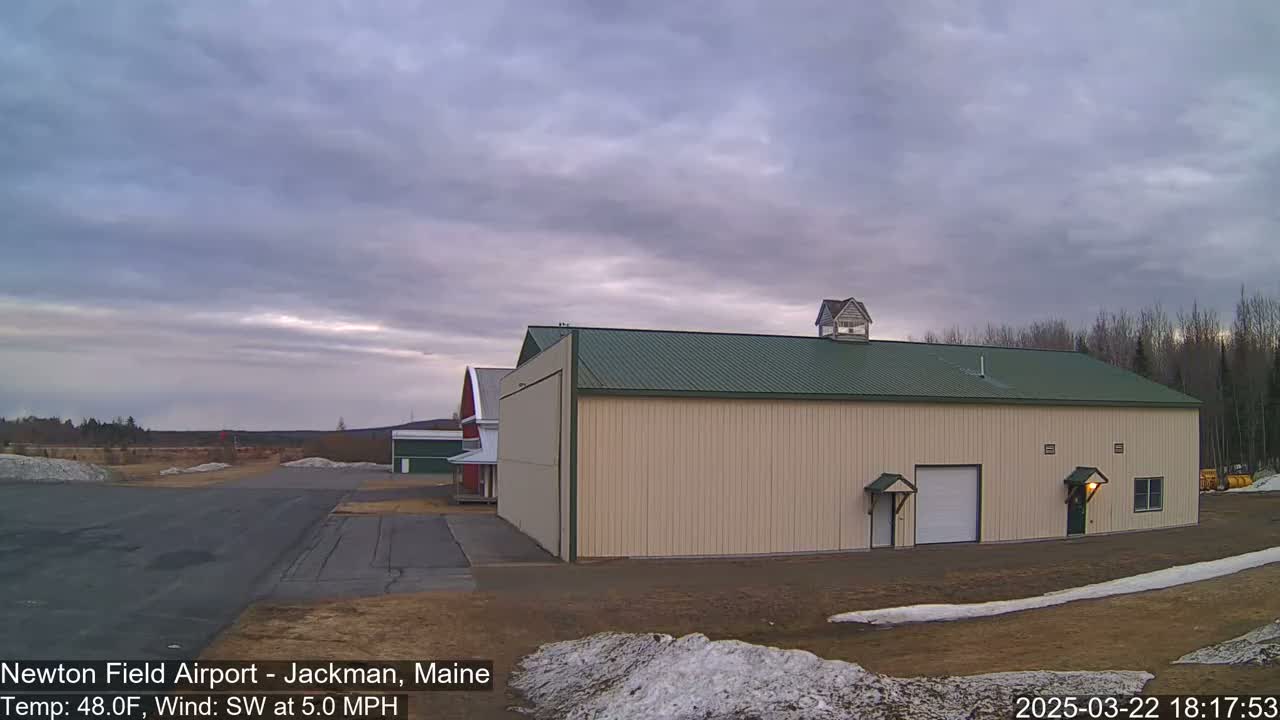 A large tan hangar with a green roof stands on an airfield under a mostly cloudy sky.