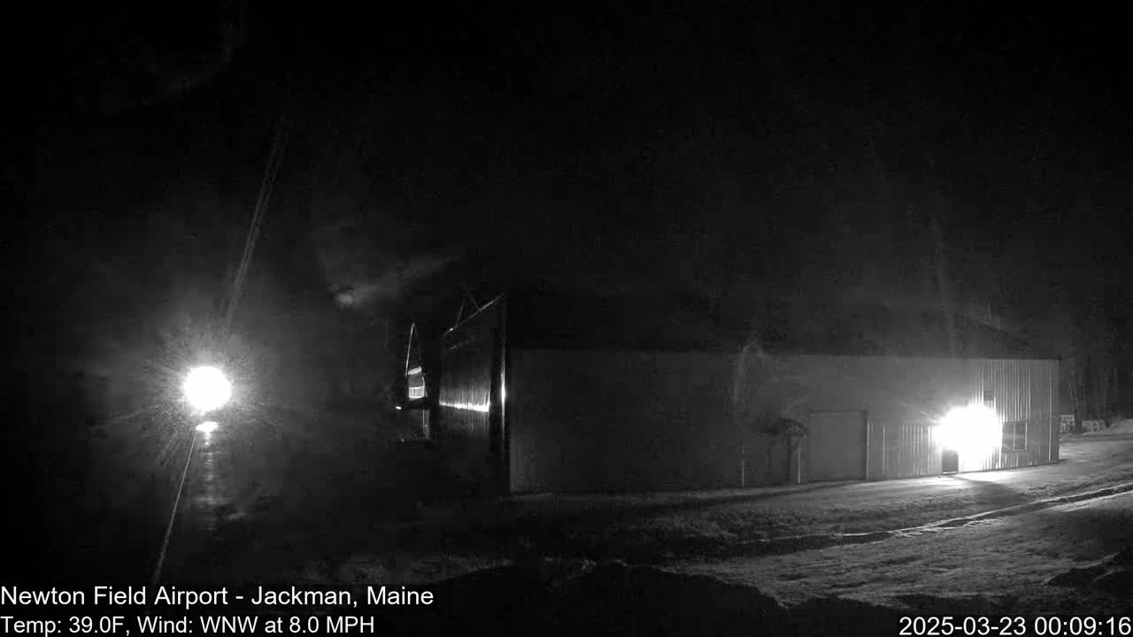 A dark, nighttime scene shows a long, low building illuminated by a bright light, with a light source visible in the distance and a slight dusting of snow on the ground under a dark sky.