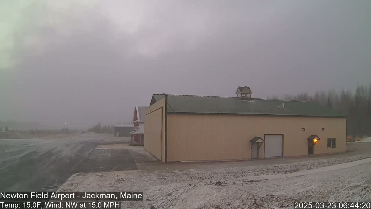 A large tan hangar stands in a snowy airfield under an overcast sky, with a light wind.