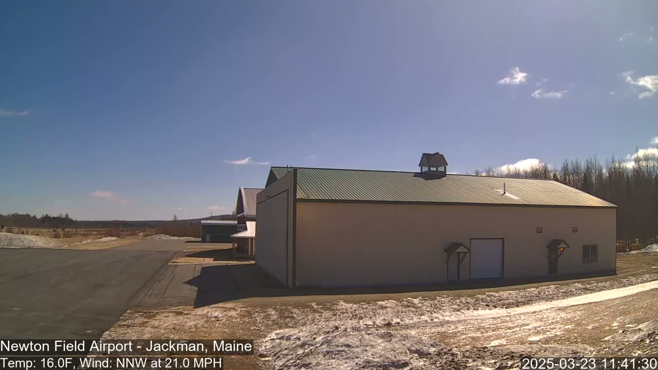 A large tan building with a green roof sits on a mostly clear day with a light wind near a paved area and sparse patches of snow.