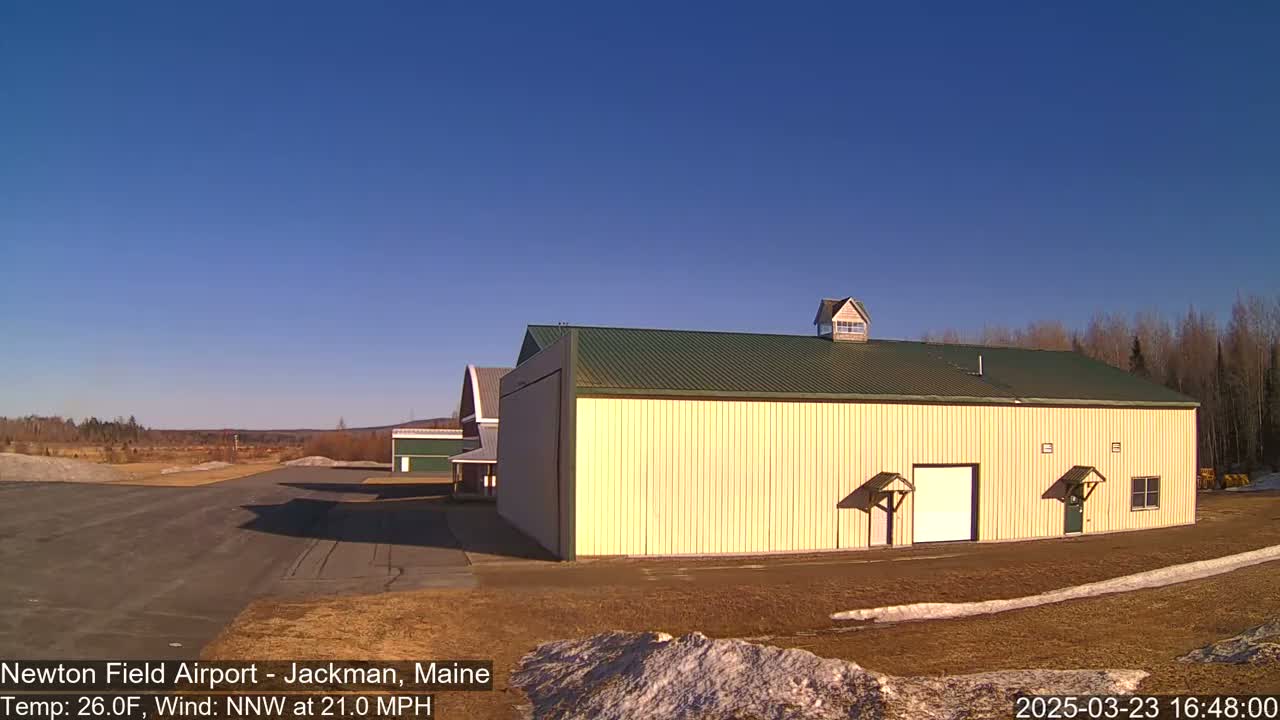 Under a clear blue sky, a large tan hangar stands beside a smaller red barn at an airfield, with a light wind blowing.