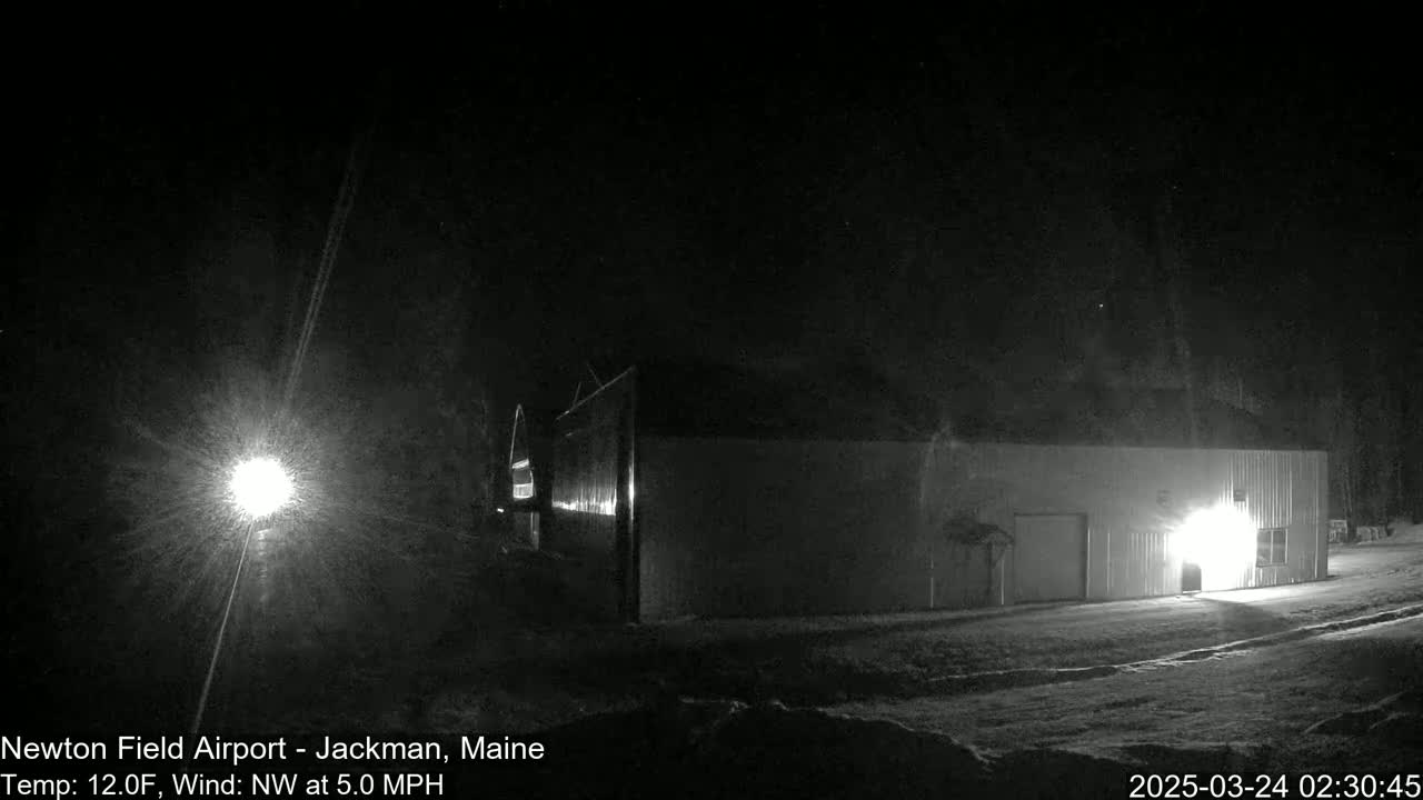 A long, low building is illuminated by lights in the snow during a nighttime snow flurry with a northwest wind.