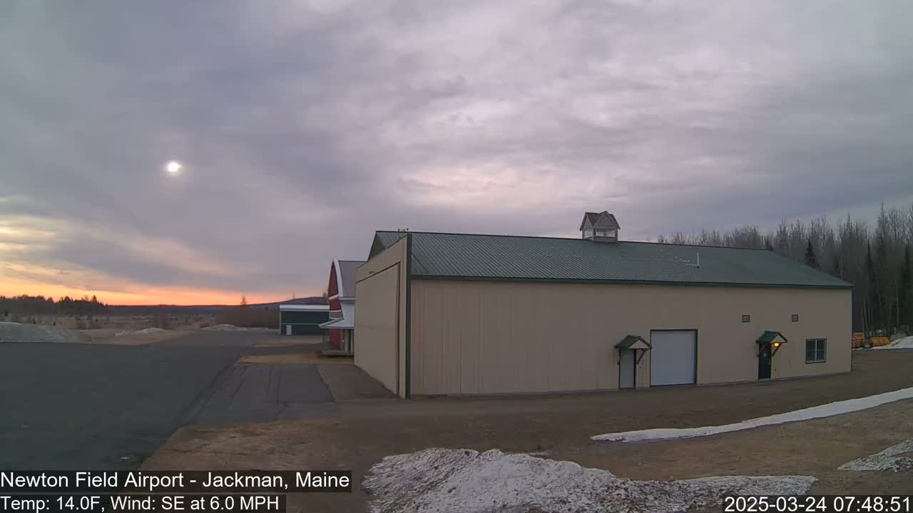 A large tan hangar with a green roof sits on an airfield under a partly cloudy sky, with a faint sunrise visible on the horizon.