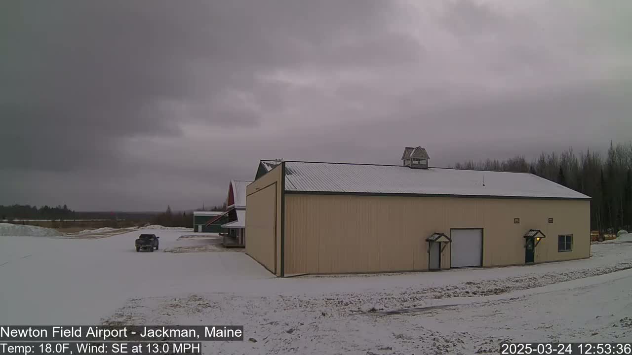 A large tan building with a snow-covered roof sits in a snowy landscape under an overcast sky, with a pickup truck parked nearby.