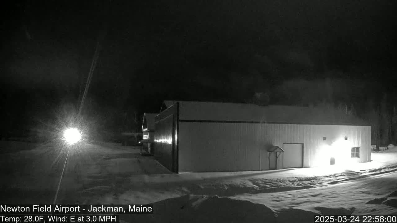 A large, light-colored building is visible at night in snowy conditions, with a light source illuminating the foreground and a faint light in the distance.