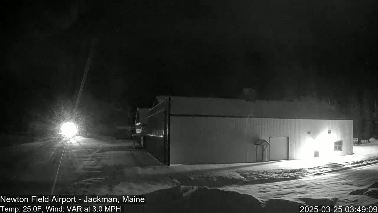 A snow-covered area at night shows a long building illuminated by a light source, with a vehicle's headlights visible in the distance.