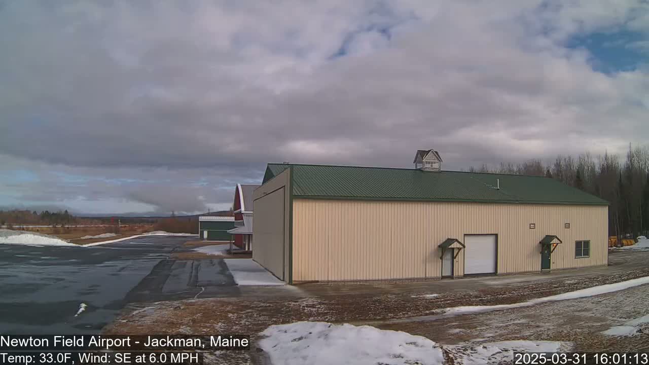 A large tan hangar with a green roof sits beside a smaller red barn on a mostly clear day with some clouds, and patches of snow on the ground.
