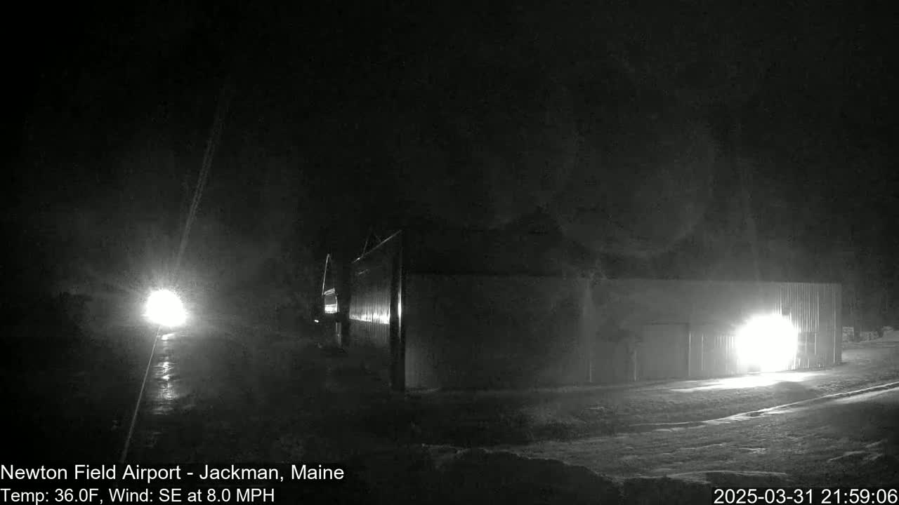 A nighttime, snow-covered scene shows a long building with lights on, illuminated by headlights of a vehicle on a road in the distance, under a dark sky with a southeast wind.