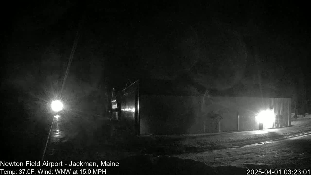 A nighttime, snowy scene shows a large building lit from within, with a bright light source visible in the distance and a 15 mph wind.
